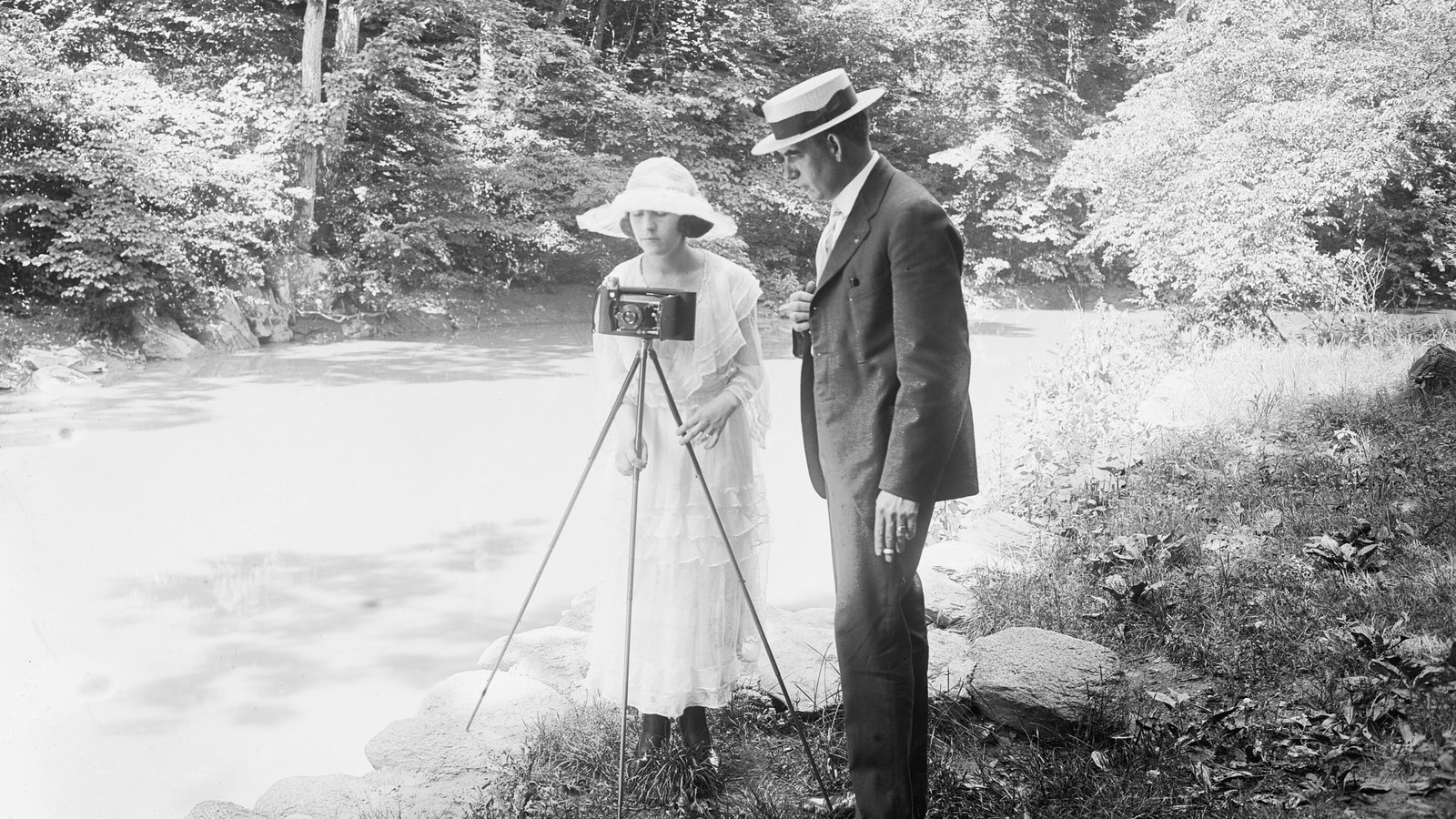 Ormanda bir dere kenarında üç ayaklı sehpaya kurulu fotoğraf makinesiyle çekim yapan bir kadın ve yanındaki adam