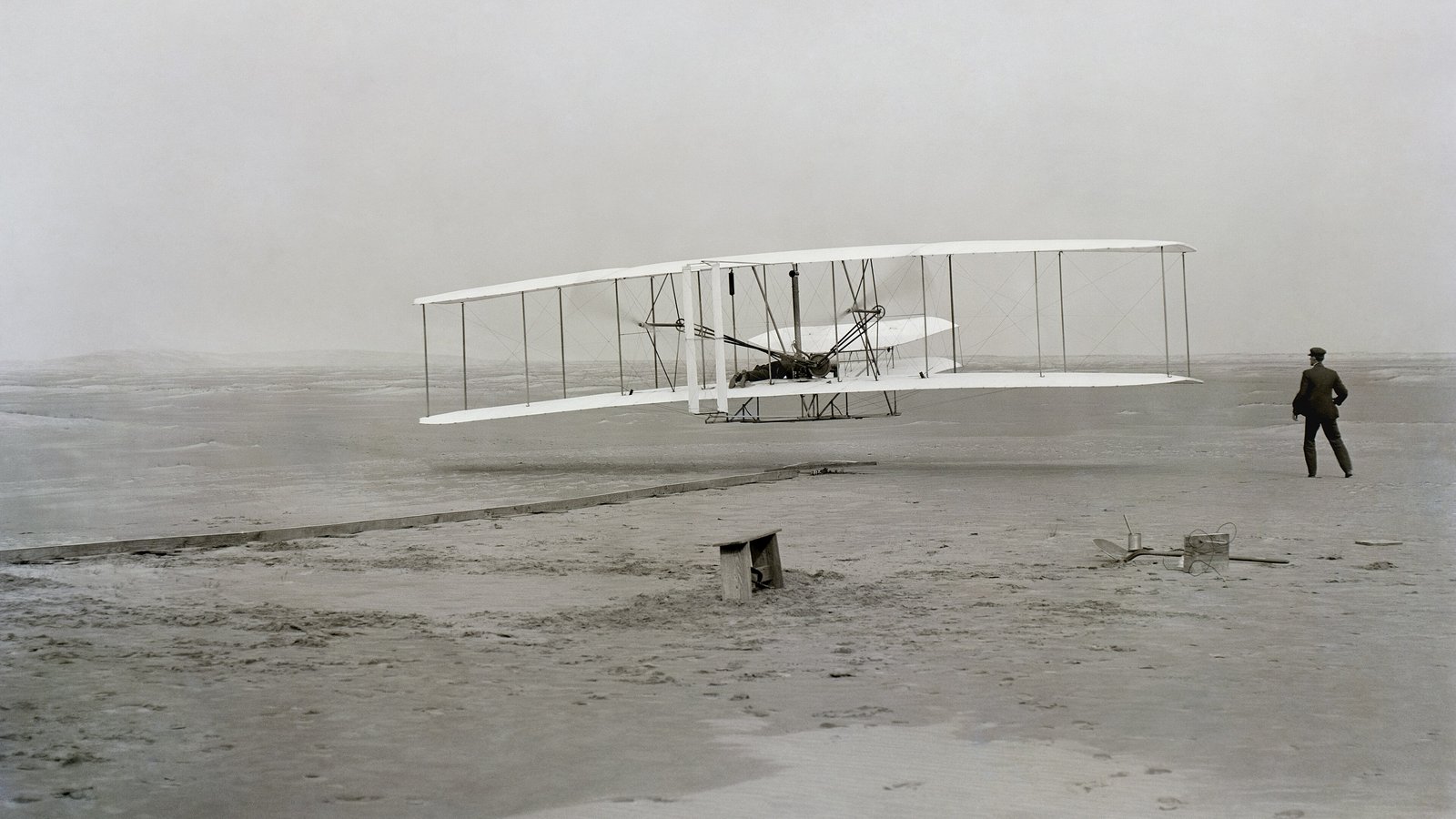 Kitty Hawk kumsalında yerden yeni kalkan çift kanatlı Wright Flyer ve yanında koşarak onu izleyen Wilbur Wright