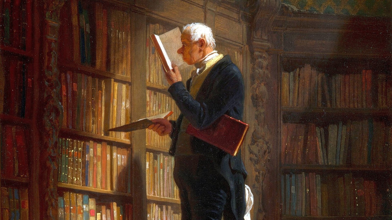 An elderly man reading a book at the top of a ladder between tall shelves in a library