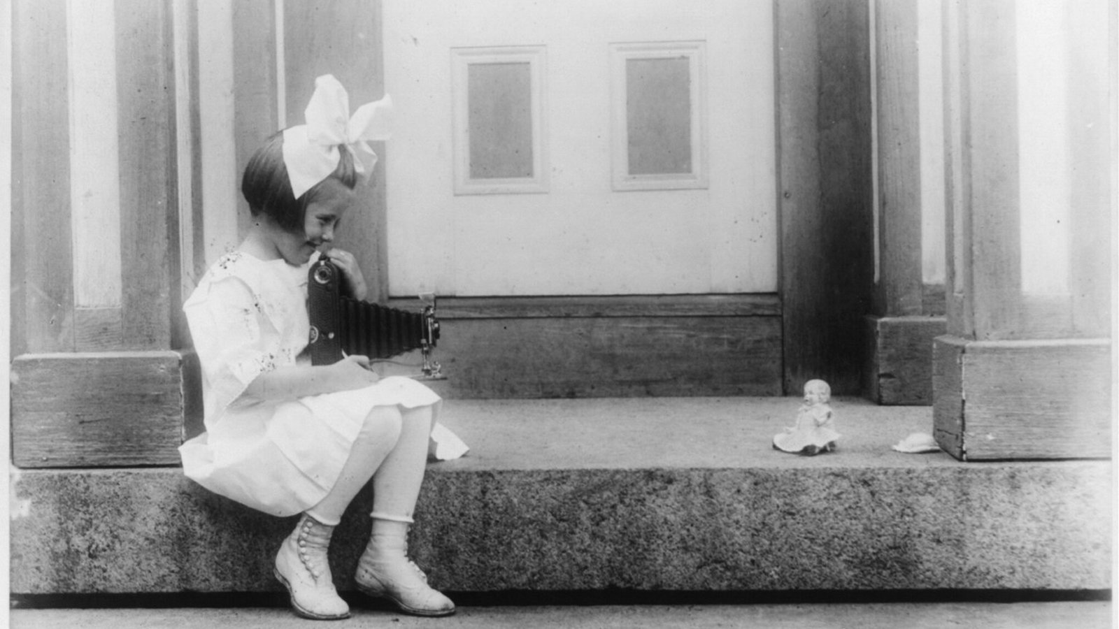 Bir evin taş basamağına oturmuş, körüklü Kodak fotoğraf makinesiyle karşısındaki oyuncak bebeğin fotoğrafını çeken küçük kız