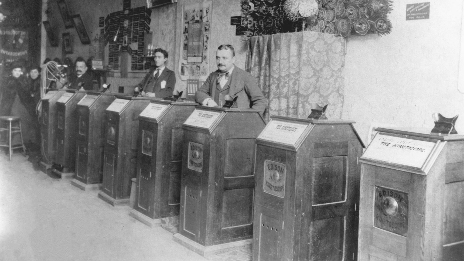 1890'larda San Francisco'da bir kinetoskop salonu: sıra hâlinde dizilmiş, tek kişinin eğilip içine baktığı ahşap Edison kinetoskop dolapları ve arkalarında duran üç adam