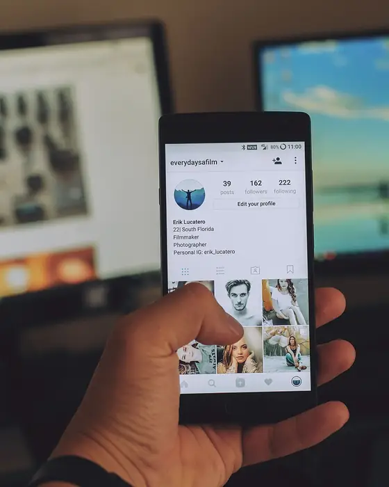 Elde tutulan telefonda açık Instagram profili, arka planda bilgisayar ekranları