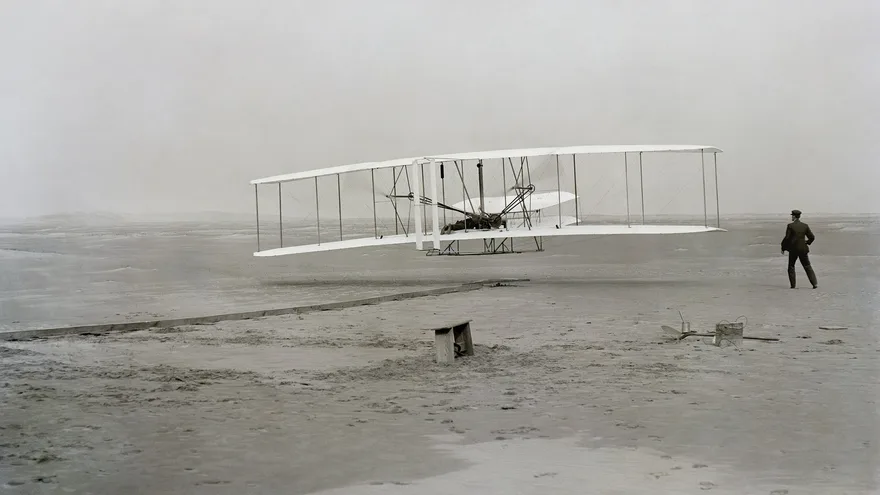 Kitty Hawk kumsalında yerden yeni kalkan çift kanatlı Wright Flyer ve yanında koşarak onu izleyen Wilbur Wright